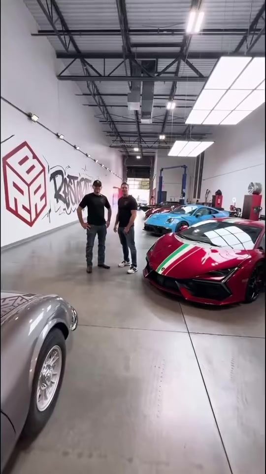 Two hosts standing among supercars in the Red Box Restoration shop